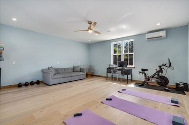 Bright multi-purpose home gym and office with light blue walls and hardwood floors, three purple yoga mats and blocks, stationary exercise bike, gray tufted sofa, small desk with computer under a window, ceiling fan and wall-mounted AC.