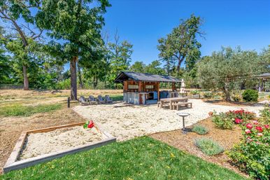 Sunny backyard oasis with a rustic covered outdoor bar/kitchen pavilion, picnic table, Adirondack chairs, sand play area and sandbox with toys, string lights, flower beds and mature trees under a clear blue sky