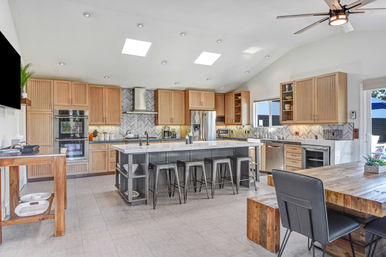 Bright open-concept kitchen with a large marble-topped island and metal bar stools, light wood cabinets, chevron tile backsplash, stainless steel appliances, skylights, ceiling fan, and rustic wood dining table.