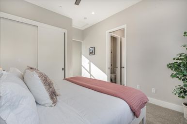 Sunlit modern master bedroom with white linens and mauve throw, sliding closet doors, open ensuite bathroom and potted plant.