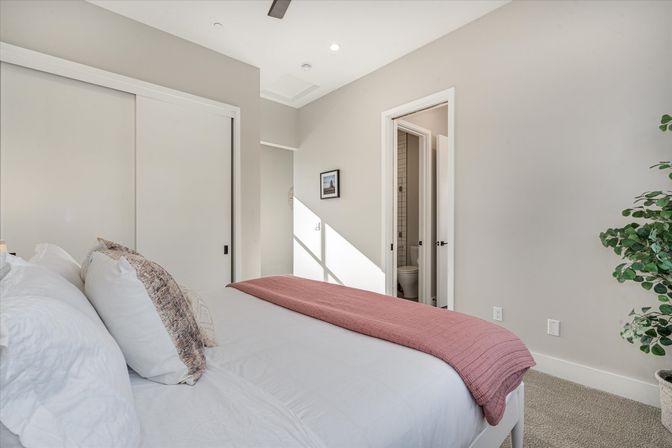 Sunlit modern master bedroom with white linens and mauve throw, sliding closet doors, open ensuite bathroom and potted plant.