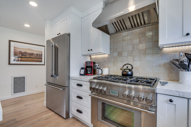Bright modern kitchen with stainless-steel French-door refrigerator and professional gas range, white shaker cabinets, tiled backsplash and hardwood floors.