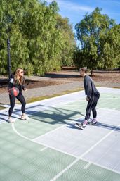 Two people in athletic wear playing on an outdoor park basketball court on a sunny day — one dribbling a red-and-black ball while the other guards near the painted free-throw area, tall green trees and blue sky in the background.