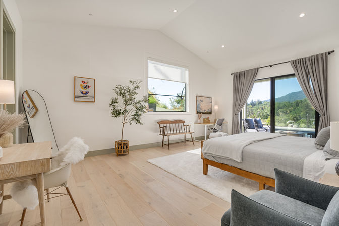 Airy modern master bedroom with light hardwood floors, king bed with gray linens, sliding glass doors to a mountain view, desk, wooden bench, potted tree and full-length mirror