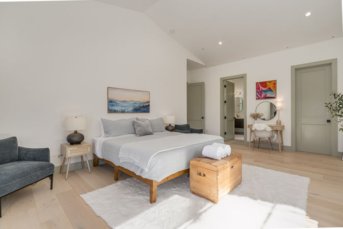 Bright modern master bedroom interior with vaulted ceiling, king bed, light wood floors, wooden trunk with towels, cozy armchair and a vanity with round mirror and colorful art