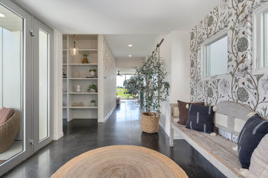 Sunlit modern entry hall with glass front doors, round jute rug on polished concrete floor, built-in white shelving, woven-basket potted tree, distressed wooden bench with dark throw pillows, and floral wallpaper leading to a bright backyard view.