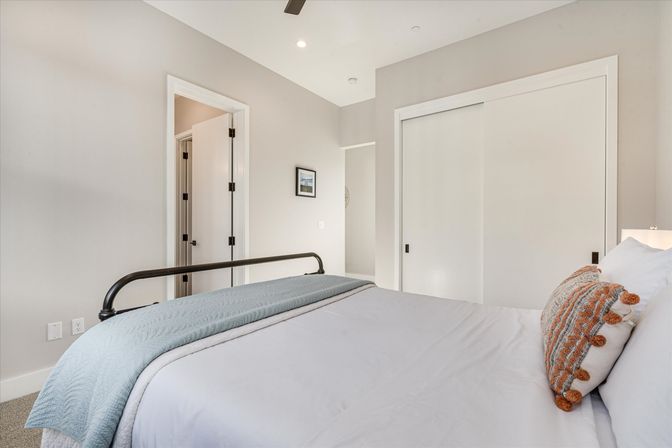 Bright modern bedroom with black metal bed frame, light-blue quilted throw, white linens and orange-tassel accent pillow, sliding closet doors and neutral-colored walls