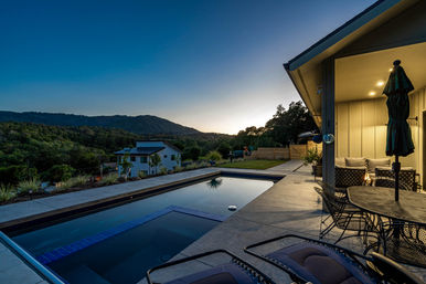 Twilight view of a modern hillside home with illuminated patio, rectangular pool, lounge chairs, outdoor dining set, and rolling mountain backdrop.