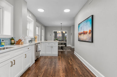 Bright modern galley kitchen with white shaker cabinets, quartz countertops, blue tile backsplash, warm hardwood floors and open-concept dining area with pendant light and framed bison painting on the wall.