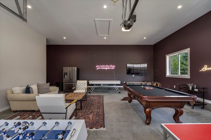 Modern residential game room with wooden pool table, foosball and ping-pong tables, air hockey, lounge seating, fridge, neon wall sign and garage-style concrete floor