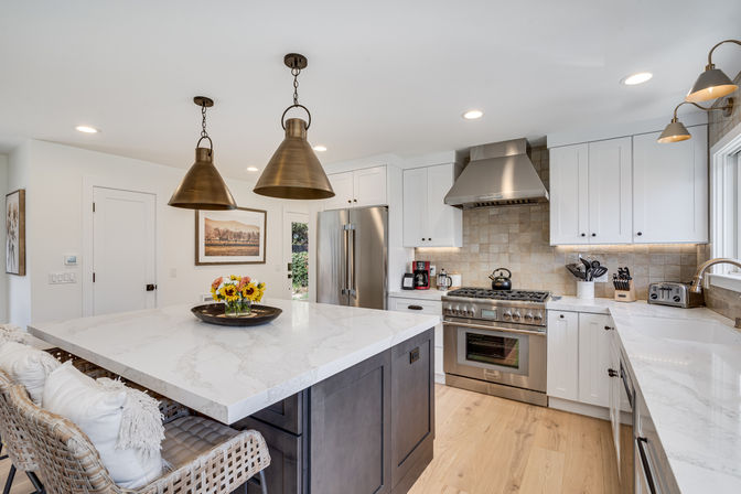 Bright, inviting modern kitchen interior with a large white marble island on a dark wood base, woven bar stools, brass pendant lights, stainless steel range and refrigerator, white shaker cabinets, neutral tile backsplash and light hardwood floors
