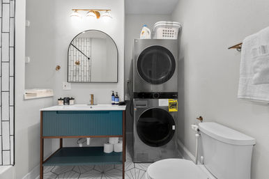 Bright modern compact bathroom-laundry with stacked stainless washer and dryer, teal vanity with arched mirror, geometric tile floor, toilet and towel rack.