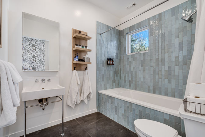 Bright modern home bathroom with blue-gray vertical subway tile tub surround, white soaking tub and shower, wall-mounted shelf with toiletries, pedestal sink and mirror, white towels and dark floor tiles.