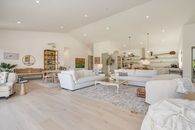 Airy sunlit open-concept living room and kitchen with vaulted ceilings, light hardwood floors, soft white sofas, rustic coffee table, dining nook and island with pendant lights.