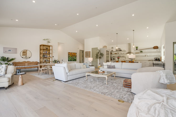 Airy sunlit open-concept living room and kitchen with vaulted ceilings, light hardwood floors, soft white sofas, rustic coffee table, dining nook and island with pendant lights.