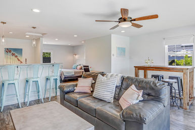 Bright open-plan coastal-style living space with a dark leather sofa and striped pillows, concrete coffee table, pale blue metal bar stools at a kitchen island, rustic wooden bar table with stools, ceiling fan, and a bed visible in the background.
