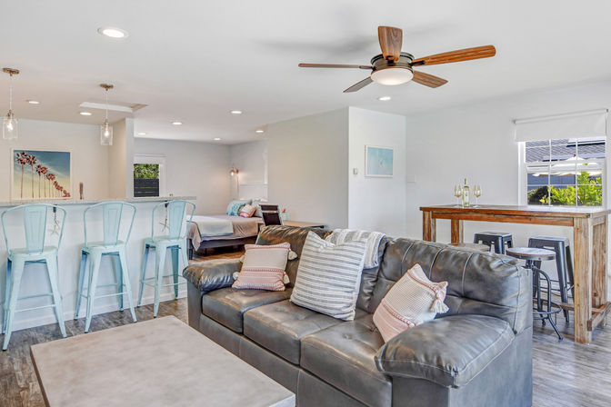 Bright open-plan coastal-style living space with a dark leather sofa and striped pillows, concrete coffee table, pale blue metal bar stools at a kitchen island, rustic wooden bar table with stools, ceiling fan, and a bed visible in the background.