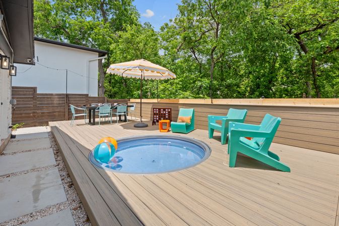 Suburban backyard wood deck with built-in round plunge pool, colorful beach balls, turquoise patio chairs, striped umbrella over al fresco dining set and leafy trees.