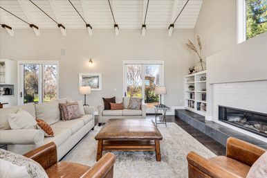 Sunlit modern farmhouse living room with vaulted white‑beamed ceiling, neutral sofas, leather ottoman, built‑in bookshelves, linear fireplace, dark hardwood floors, and sliding glass doors to a yard.