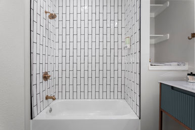 Sleek tub-shower with white vertical subway tiles and black grout, brass shower fixtures and wall-mounted soap dispenser, white bathtub and blue vanity with open shelving — modern bathroom design.