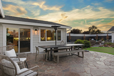 Inviting suburban backyard patio at sunset with stone flooring, a long wooden picnic table and benches, wicker lounge chairs, patio heater, small putting green and landscaped shrub borders.