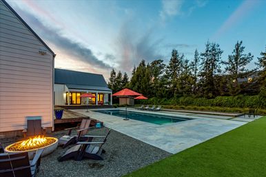 Modern backyard at dusk with a rectangular swimming pool, concrete patio, red umbrellas over lounge chairs, a glowing fire pit surrounded by Adirondack chairs, and a farmhouse-style house with tall trees in the background.