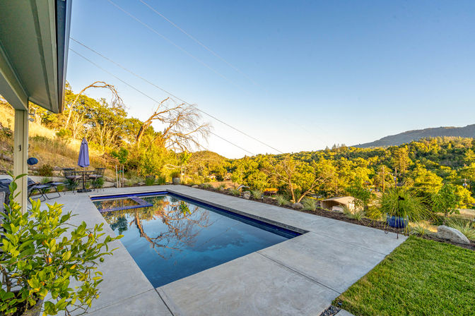 Rectangular backyard pool on a concrete patio reflecting trees and clear blue sky, with lounge chairs, umbrella and potted plants overlooking sunlit hillside and distant mountain views during golden hour.