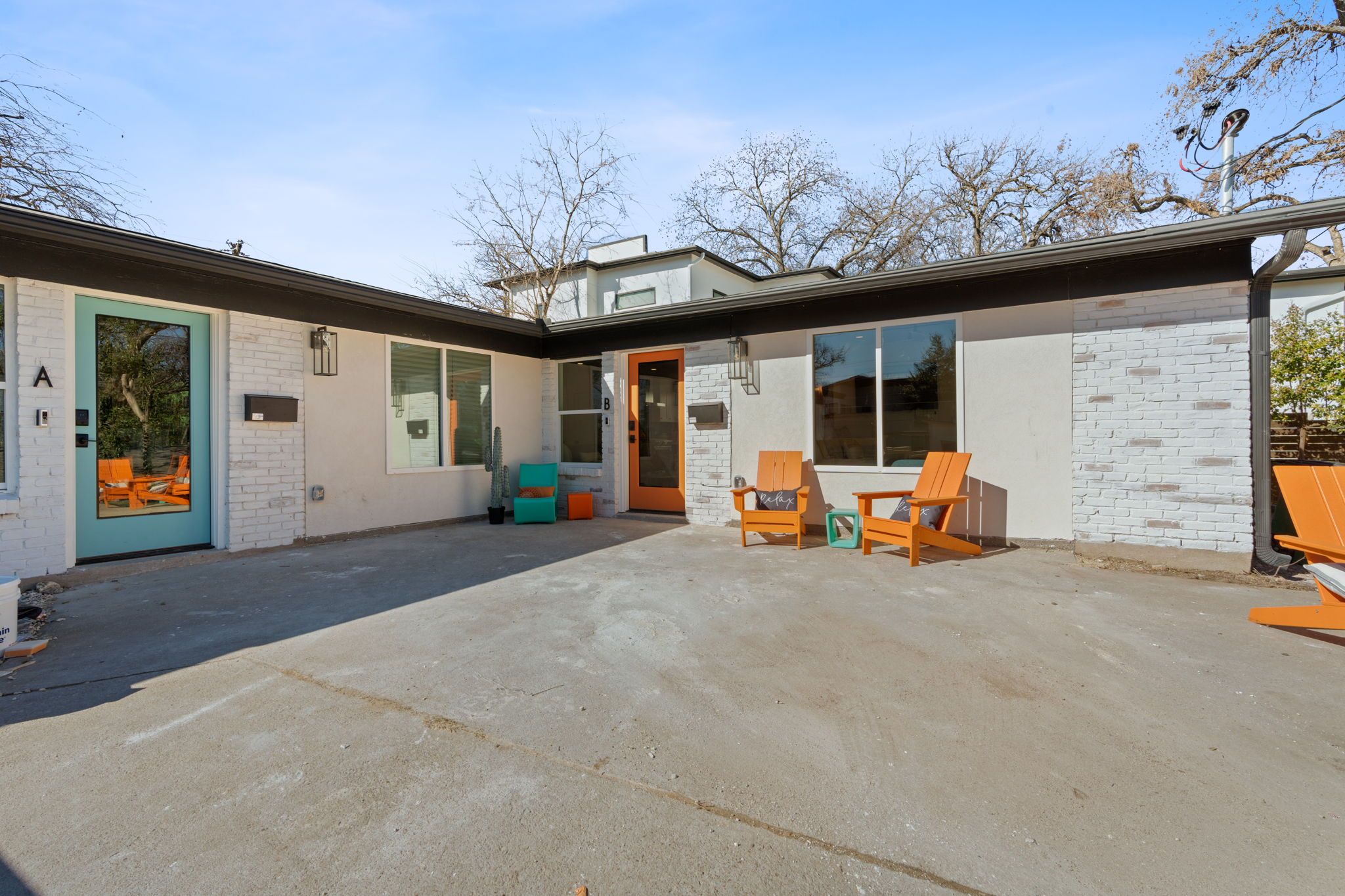 Sunlit mid-century modern duplex courtyard with teal and orange front doors, white brick exterior, concrete patio, and bright orange Adirondack chairs