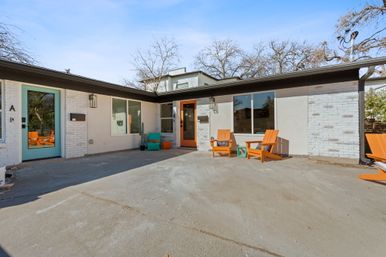 Sunlit mid-century modern duplex courtyard with teal and orange front doors, white brick exterior, concrete patio, and bright orange Adirondack chairs