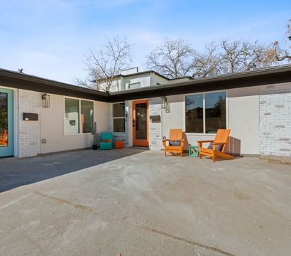 Sunlit mid-century modern duplex courtyard with teal and orange front doors, white brick exterior, concrete patio, and bright orange Adirondack chairs