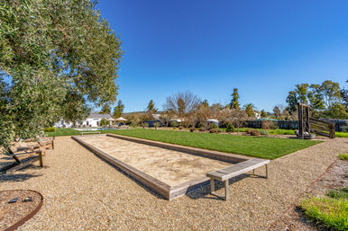 Sunlit backyard bocce court on gravel with wooden benches, manicured lawn, trees and a clear blue sky