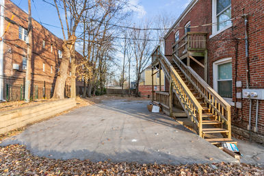 Urban residential backyard with concrete pad and wooden exterior staircase to a second-floor entrance of a red brick rowhouse, leaf-strewn ground, bare trees, neighboring brick buildings and utility meters under a clear blue sky.