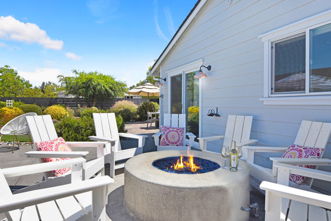 Sunny suburban backyard patio with white Adirondack chairs and pink patterned cushions arranged around a round concrete fire pit with blue glass rocks and a small flame; bottle of white wine and two glasses on the pit edge, sliding glass doors on a light-gray house, landscaped garden and umbrella in the background.