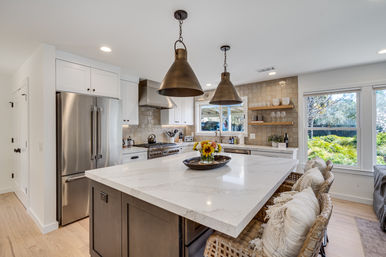 Bright modern open-concept kitchen with large white marble island, bronze pendant lights, stainless-steel appliances, tile backsplash, wicker bar stools, and windows overlooking a green backyard.
