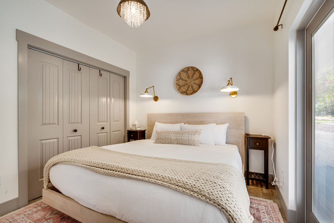 Cozy modern farmhouse bedroom with king-size bed dressed in white linens and a chunky cream knit throw, neutral wood headboard, brass wall sconces, woven wall decor, matching dark wood nightstands, sliding closet doors and a glass door flooding the room with natural light.
