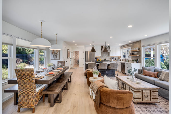 Sunlit open-plan modern farmhouse interior with a wooden dining table and wicker chairs, cozy living area, and island kitchen with bar stools overlooking a garden