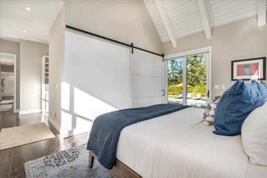 Bright modern bedroom with vaulted white-beamed ceiling, hardwood floors, sliding white barn door, navy quilted throw on the bed and floor-to-ceiling glass doors revealing a backyard pool and patio umbrella.