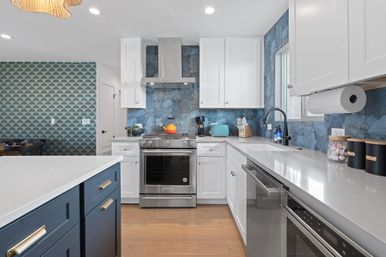 Modern coastal-inspired kitchen with blue patterned tile backsplash and scallop accent wall, white shaker cabinets, navy island with brass pulls, stainless steel range and hood, quartz countertops, hardwood floors and pops of teal and orange.