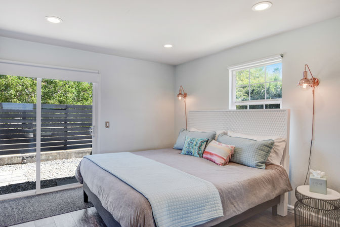 Bright modern bedroom with king-size bed, light-blue quilt and colorful throw pillows, white textured headboard, copper wall sconces, sliding glass door to a fenced patio and small window showing greenery.