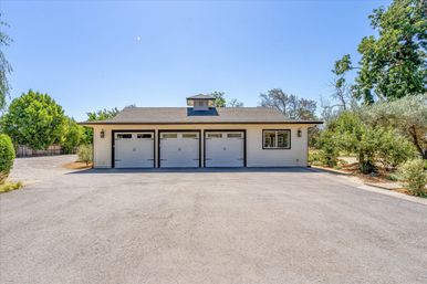 Three-car detached garage with white siding and black trim, rooftop cupola, wide asphalt driveway and trees under a clear blue sky