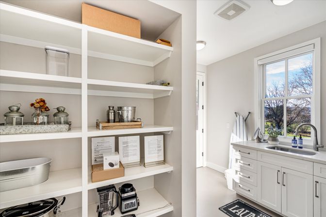 Sunny modern laundry/mudroom in a residential home with built-in white shelves holding metal containers, small appliances and decor, next to white shaker cabinets, a stainless sink and a window with a yard view.