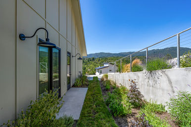 Narrow side yard of a modern hillside home with board-and-batten siding, black gooseneck wall lights, concrete path and planted borders opening to mountain views under a clear blue sky.