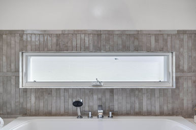 Spa-like modern bathtub with chrome faucet and handheld shower beneath a slim frosted horizontal window set in narrow vertical gray tile backsplash.