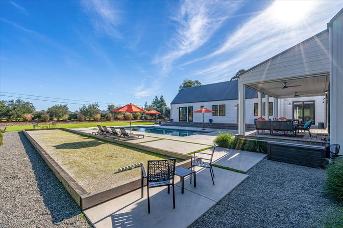 Sunlit modern farmhouse backyard with rectangular pool, lounge chairs and red umbrellas, covered patio dining area, and a gravel bocce-style court under a bright blue sky.