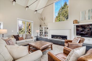 Sunlit modern living room with vaulted white ceiling and tall window, neutral sofas, leather armchairs, oversized leather ottoman, built-in bookshelves and a linear fireplace.