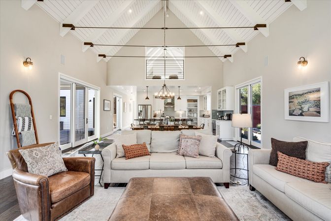 Bright open-concept living room with vaulted white-beamed ceiling, neutral linen sofas, leather armchair and large ottoman, sliding glass doors, chandelier-lit open kitchen and dining area in modern farmhouse style.