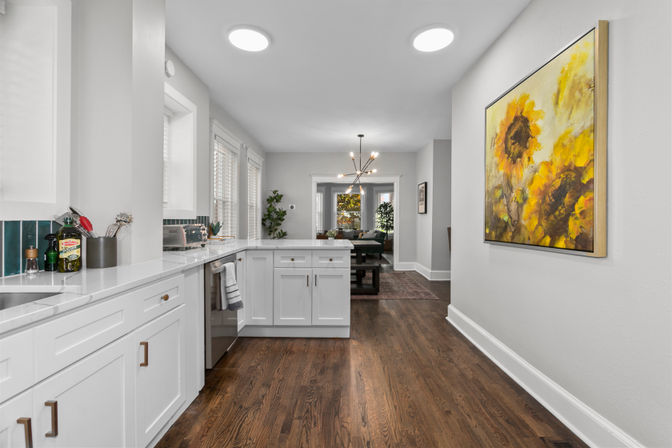 Bright modern open-concept kitchen with white shaker cabinets, marble countertops, green tile backsplash and hardwood floors; peninsula opens to a living room with a mid-century chandelier and large sunflower wall art.