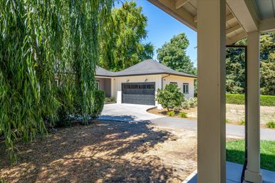 Sunny suburban driveway curving to a detached two-car garage with a dark paneled door and light siding, framed by a weeping willow, mature trees, and shaded porch posts under a clear blue sky.