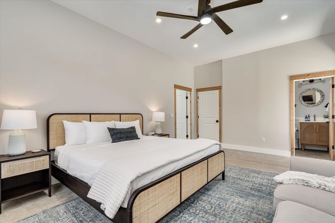 Modern neutral master bedroom with rattan-framed king bed, white bedding, matching nightstands and lamps, blue patterned area rug, ceiling fan, and open doorway to an en-suite bathroom