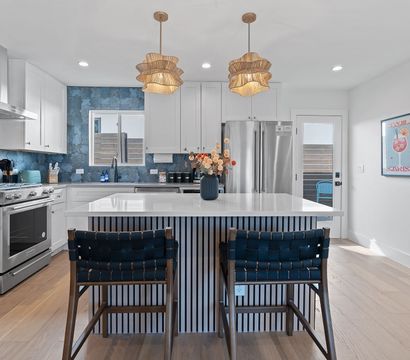 Bright contemporary white kitchen with large marble-topped island, blue patterned tile backsplash, stainless steel range and refrigerator, woven navy bar stools, rattan pendant lights, hardwood floors and a floral vase centerpiece.